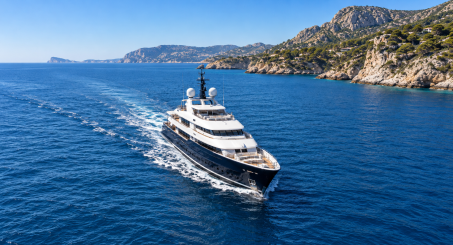 White superyacht with a dark hull cruising through blue water beside a rocky coastline.